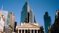 Historic Royal Exchange building in London with modern skyscrapers and clear blue sky overhead.