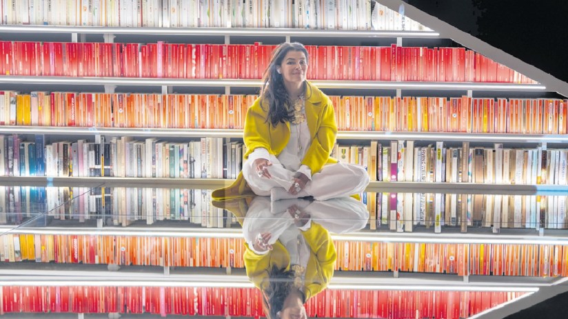 Es Devlin, a woman in a yellow coat, sits cross-legged on a mirrored floor in front of bookshelves
