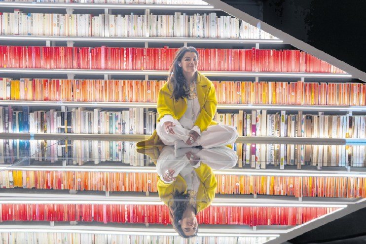 Es Devlin, a woman in a yellow coat, sits cross-legged on a mirrored floor in front of bookshelves