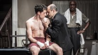 A boxing manager in a suit talks to a shirtless boxer with bandaged hands, while another man watches in the background.