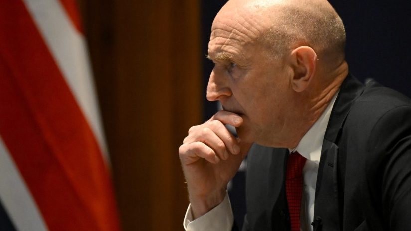 Patrick Healey in a suit with a red tie, looking contemplatively to the left, with an American flag behind him.