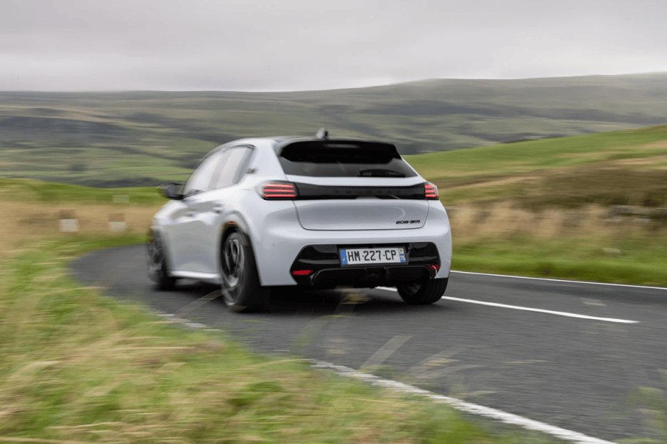 White Peugeot 208 GTi hatchback speeding on a winding road with blurred green hills in the background.