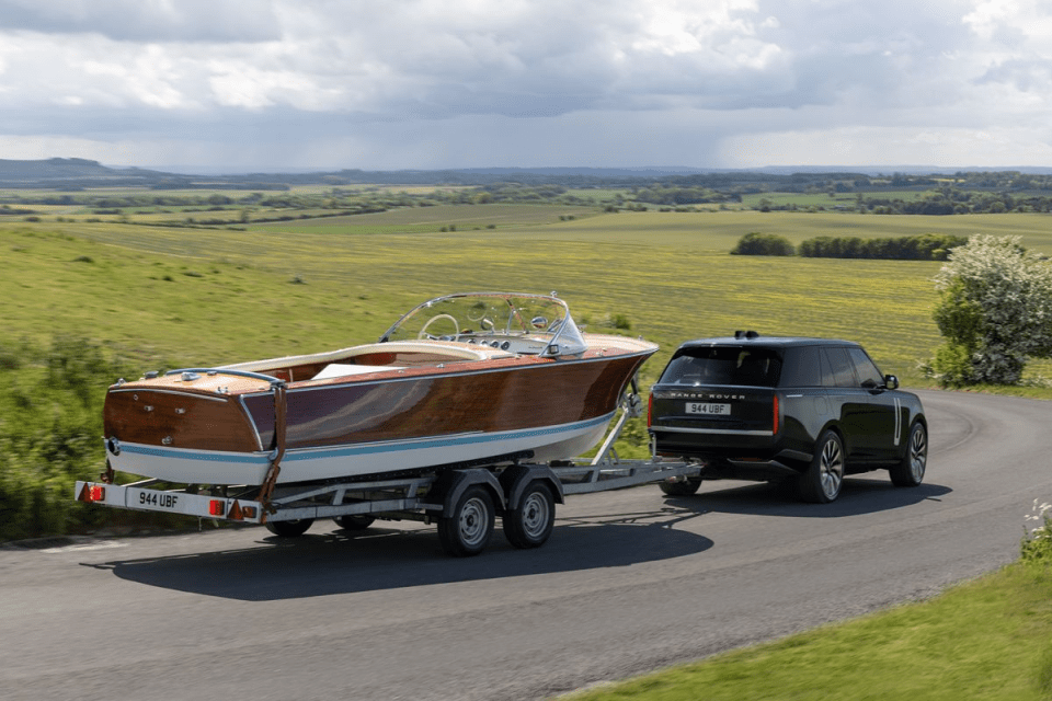 Dark green Range Rover towing a classic wooden boat on a trailer down a winding road through green fields.