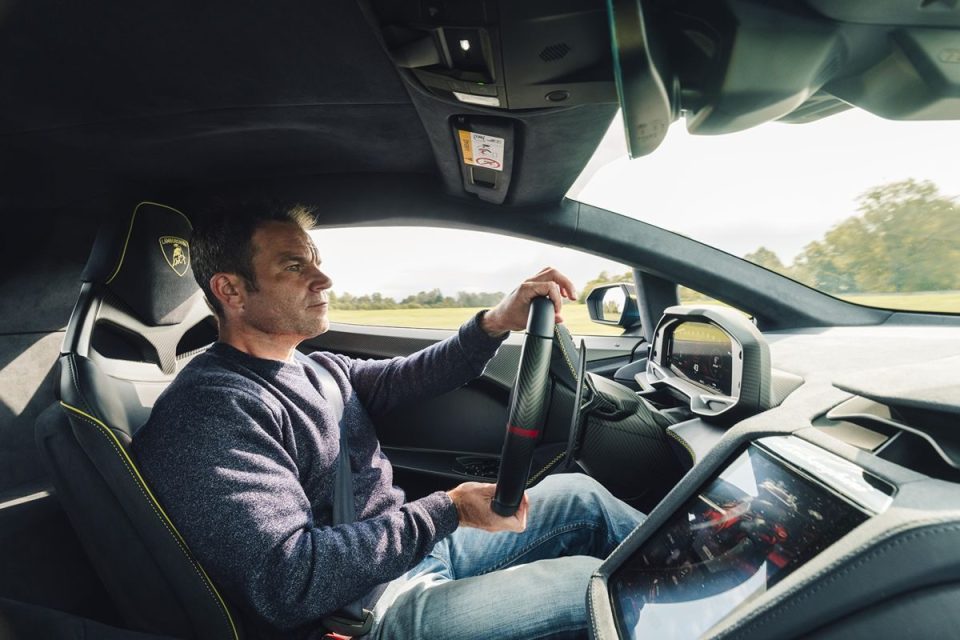 Man in a gray sweater and jeans driving a luxury sports car, holding the steering wheel.