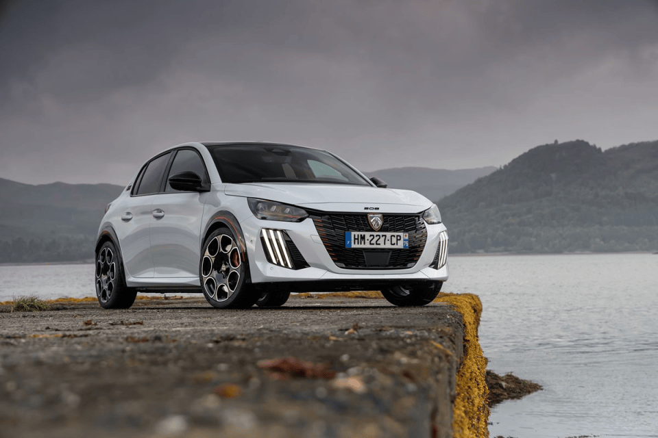 White Peugeot 208 parked on a concrete pier overlooking a lake with mountains under a cloudy sky.
