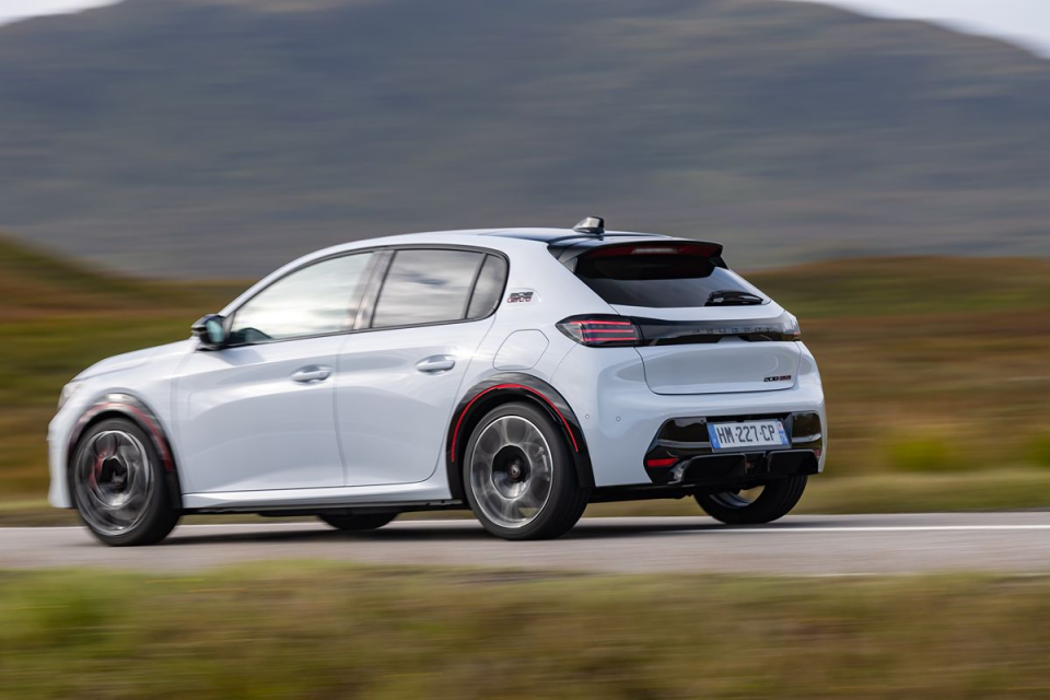White Peugeot 208 GT-Line driving on a road with blurred background, showcasing its sporty design and taillights.