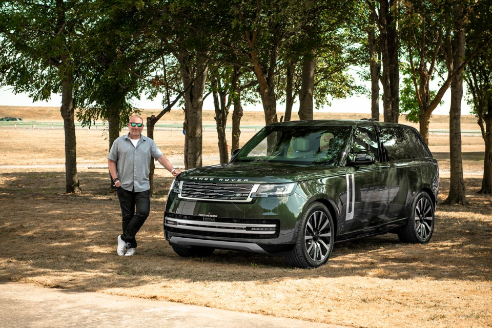 Man in sunglasses and gray shirt leaning on a dark green Range Rover SUV, outdoors with trees.