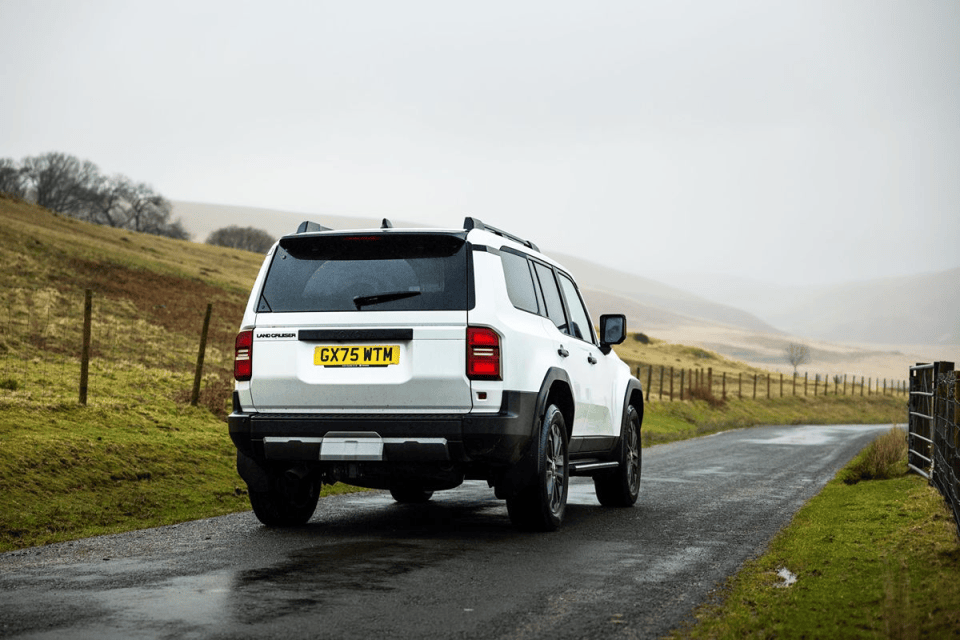 White Toyota Land Cruiser SUV driving on a wet country road with rolling hills in the background under a cloudy sky.