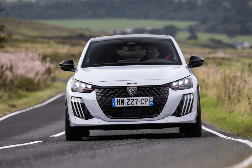 White Peugeot 208 driving on a winding country road, front view, with a driver in sunglasses visible.