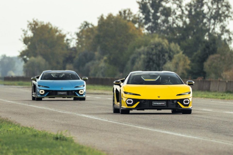 Two sleek supercars, one yellow and one blue, driving on a track with trees in the background.