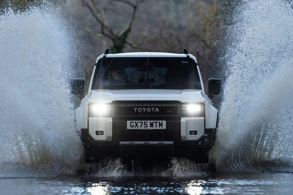 White Toyota SUV driving through a large puddle, splashing water high on both sides, headlights on.