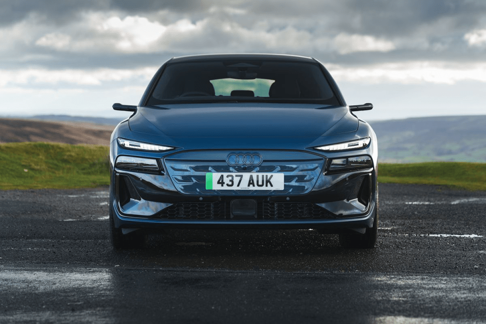 Front view of a dark blue Audi electric car on a wet road with a cloudy sky and green hills in the background.