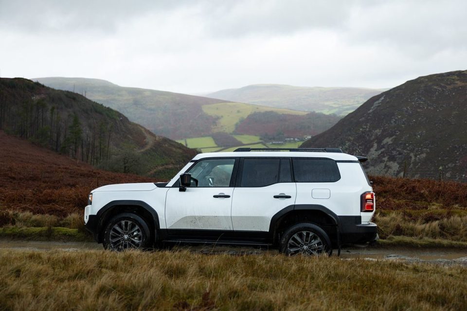 White SUV on a muddy track with rolling green and brown hills in the background under a cloudy sky