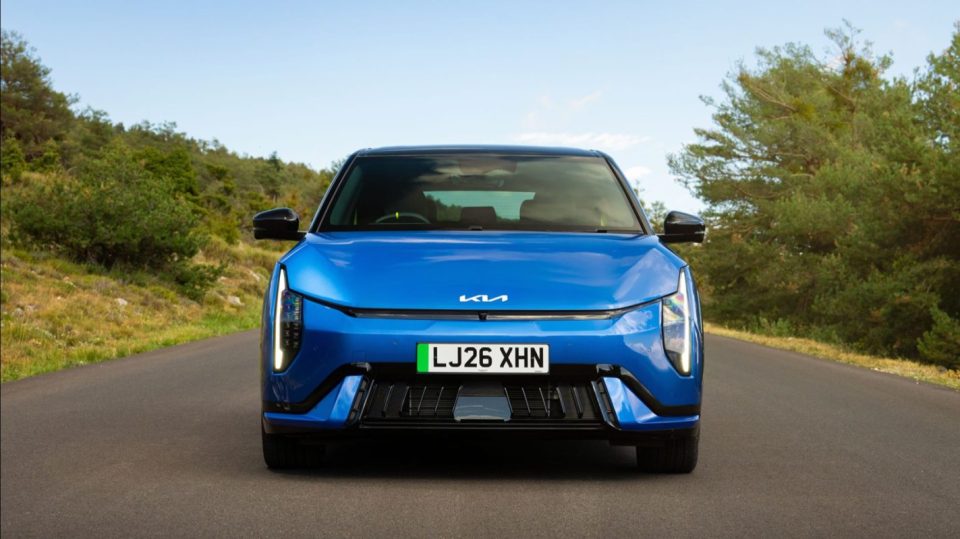 Front view of a blue Kia EV6 electric car on an asphalt road with trees and hills in the background.