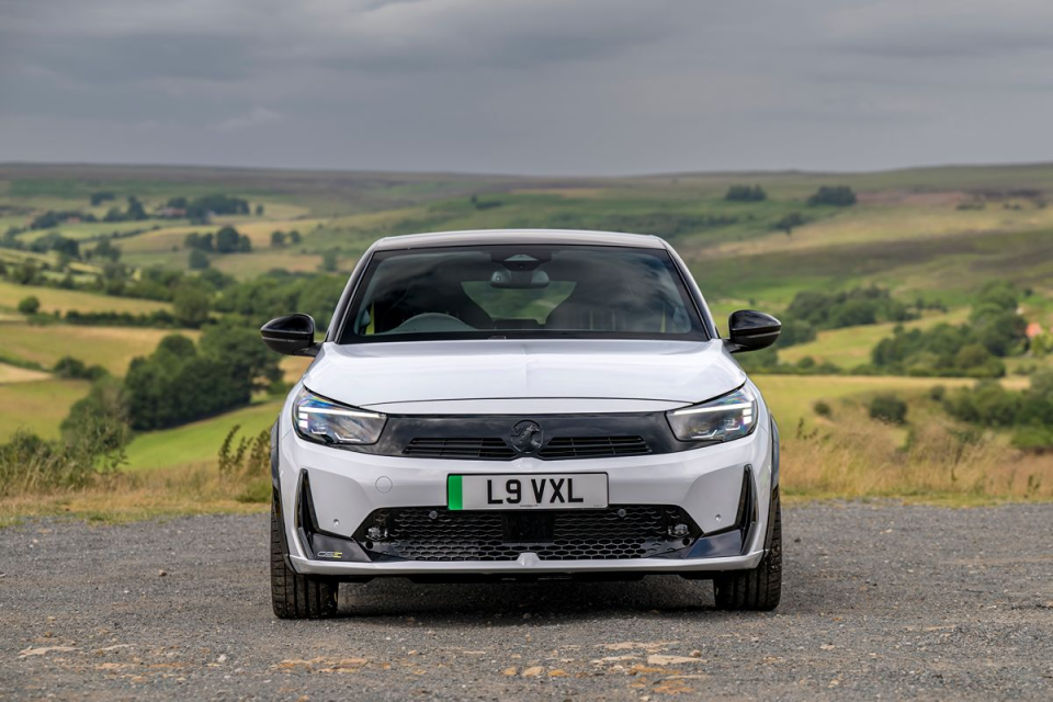 Front view of a white Vauxhall Corsa parked on a gravel road with rolling green hills and a cloudy sky in the background.