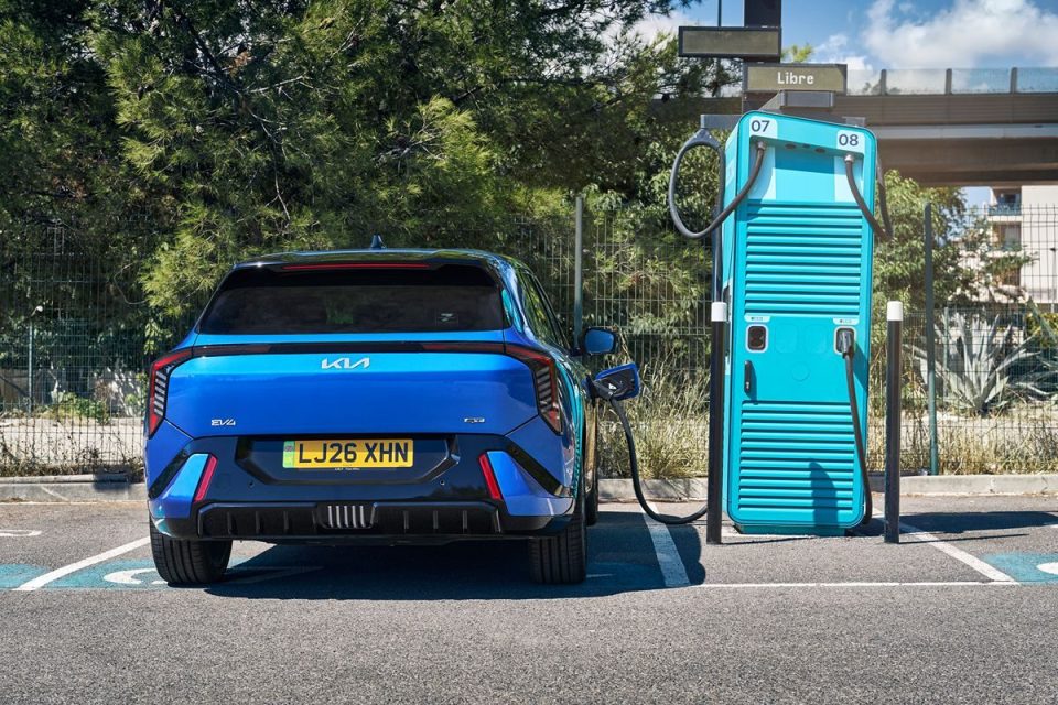 Blue Kia EV6 electric vehicle charging at a turquoise public charging station with green trees in the background.