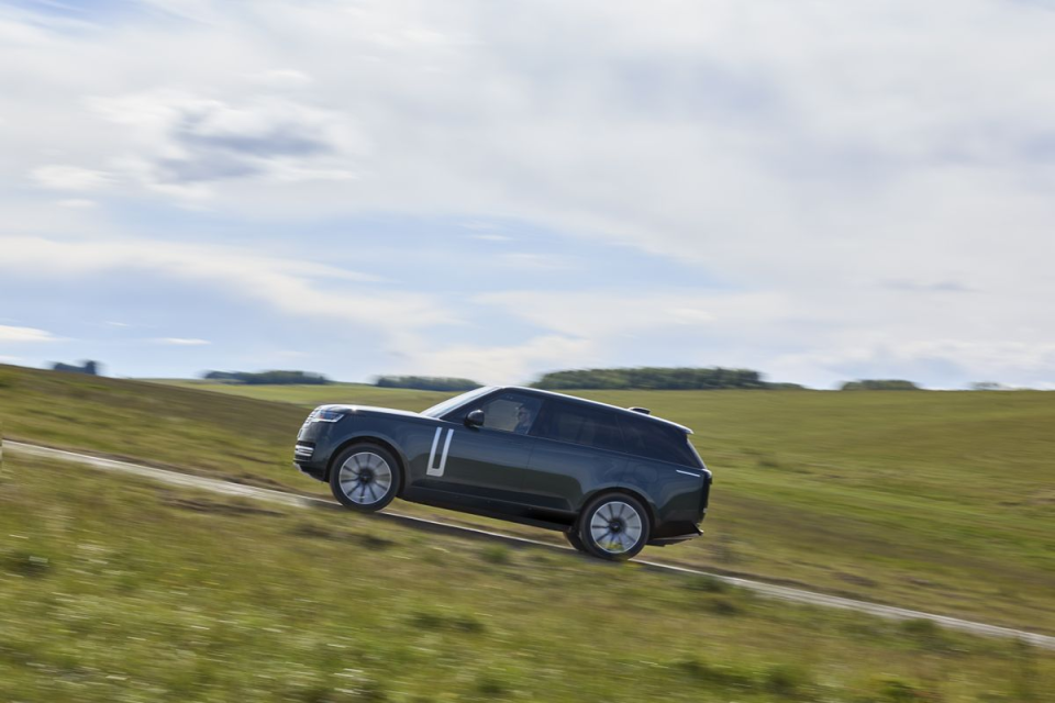 Dark gray Range Rover SUV driving uphill on a winding road through a green, grassy landscape under a cloudy sky.