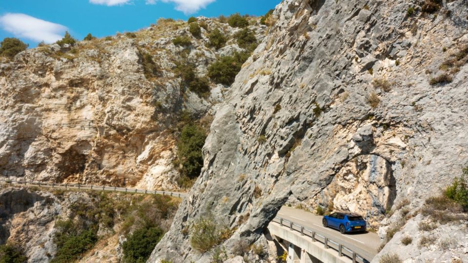 Blue SUV driving on a winding mountain road built into a rugged rock face under a clear sky.