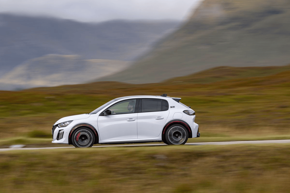 White Peugeot 208 hatchback driving on a rural road with motion blur, rugged hills in the background.