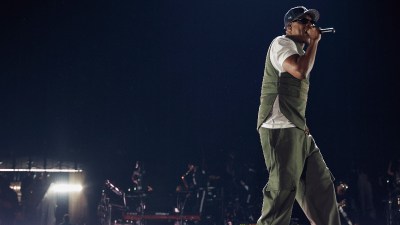 Jay-Z performing on stage at Tottenham Hotspur Stadium, wearing a cap, sunglasses, vest, and cargo pants.