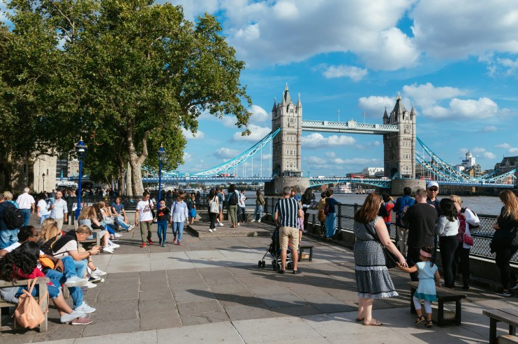 Crowd of tourists and locals walking along the Thames River with the iconic Tower Bridge in London, UK, on a sunny day.
