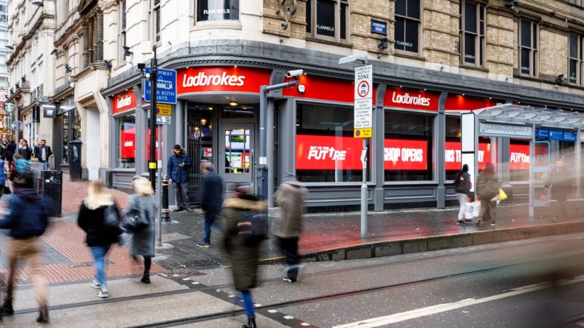 Exterior view of a Ladbrokes betting shop with red signage on a busy city street, pedestrians walking by.