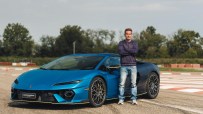 Man in a blue sweater and jeans standing beside a blue Lamborghini on a racetrack.