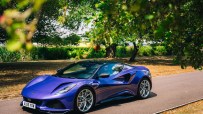 Purple Lotus Emira sports car parked on an asphalt road under green trees on a sunny day
