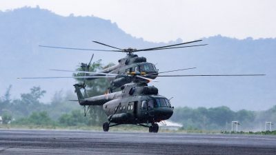Two military helicopters, dark green, preparing for takeoff on a wet runway with misty mountains in the background