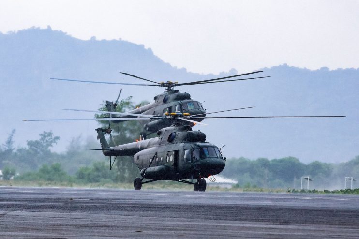Two military helicopters, dark green, preparing for takeoff on a wet runway with misty mountains in the background