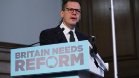 Robert Jenrick speaking at a podium with BRITAIN NEEDS REFORM sign, wearing glasses, suit, and green tie.