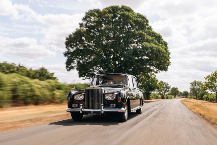 Classic black Rolls-Royce sedan driving on a country road with blurred background foliage and a large tree.