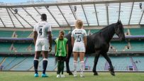 Maro Itoje, Lark Davies, and a child stand with a black horse on a rugby pitch, facing empty stadium seats.
