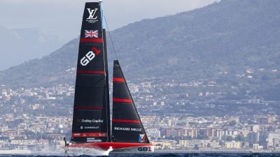 Racing sailboat GB1 with sponsors Oakley Capital and Richard Mille, flying British flag, against a coastal city and mountains