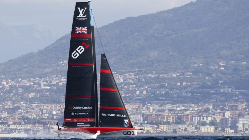 Racing sailboat GB1 with sponsors Oakley Capital and Richard Mille, flying British flag, against a coastal city and mountains