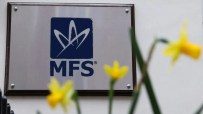 MFS logo on a silver plaque with a brown border, mounted on a white wall, blurred yellow flowers in foreground