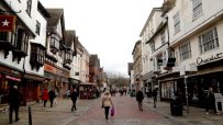 Pedestrianized street with historic buildings, shops like Hotel Chocolat and Flight Centre, and people walking.