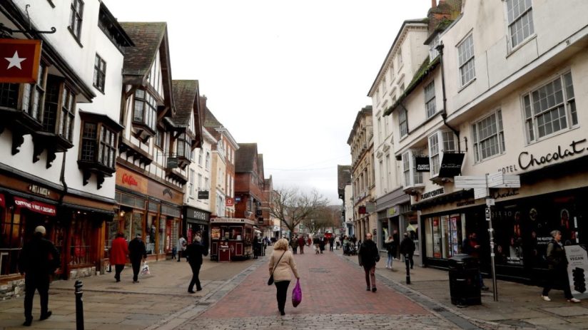 Pedestrianized street with historic buildings, shops like Hotel Chocolat and Flight Centre, and people walking.