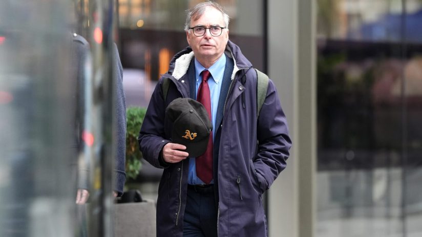 Man in glasses wearing a blue shirt, red tie, navy coat, and holding a black baseball cap outdoors.