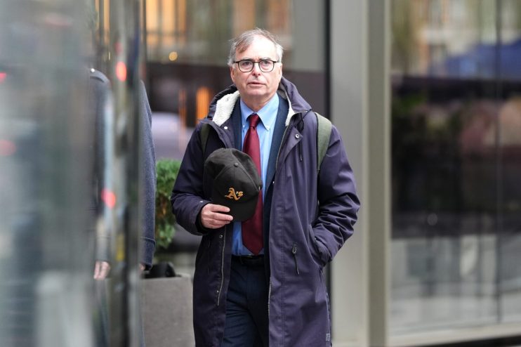 Man in glasses wearing a blue shirt, red tie, navy coat, and holding a black baseball cap outdoors.