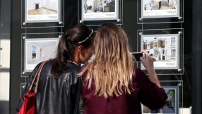 Two women view real estate listings in a Parkheath agency window, one taking a photo with her phone.