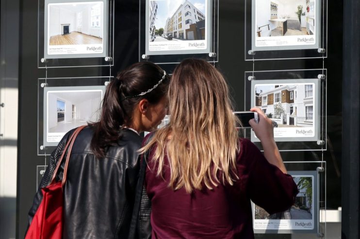 Two women view real estate listings in a Parkheath agency window, one taking a photo with her phone.