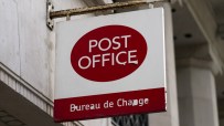 Post Office and Bureau de Change sign, white with red lettering, hanging from a rusted bracket on a building.
