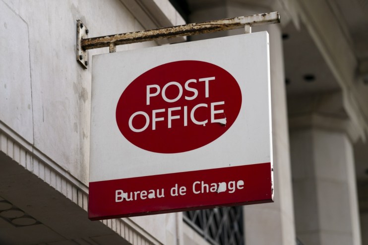 Post Office and Bureau de Change sign, white with red lettering, hanging from a rusted bracket on a building.