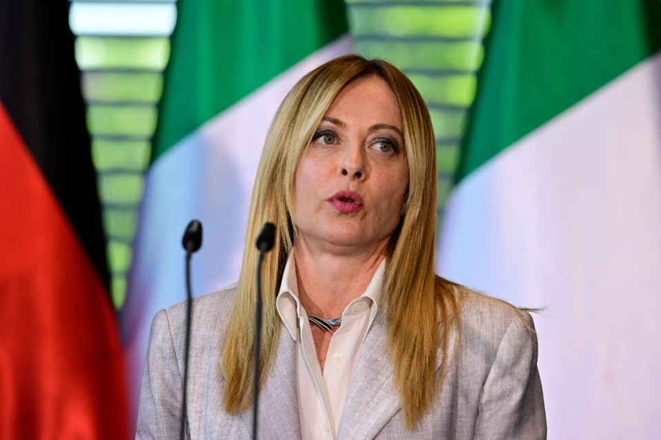 Giorgia Meloni speaking at a podium with microphones, Italian flag in background