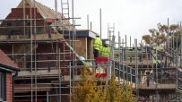 Construction workers in hard hats and high-vis jackets on scaffolding, building a new brick house with roof timbers.
