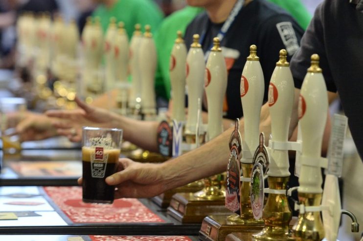 Person holding a glass of dark beer near multiple beer taps at a brewery or bar.