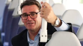 Andy Burnham smiling on a bus or train, holding a white grab handle, discussing new transport fines.