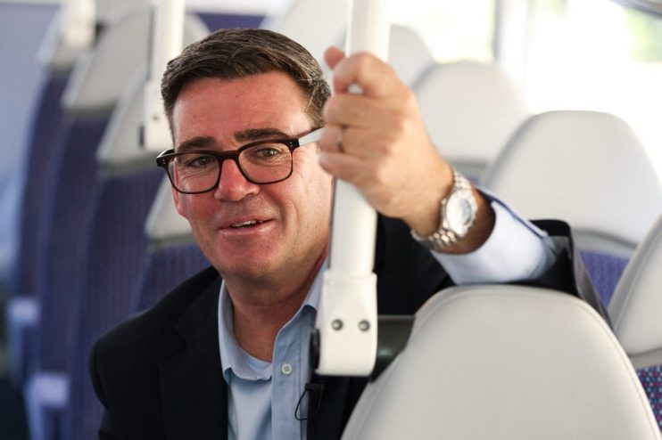 Andy Burnham smiling on a bus or train, holding a white grab handle, discussing new transport fines.