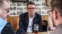 Andy Burnham, Mayor of Greater Manchester, in a dark suit and glasses, sitting at a table with a beer.